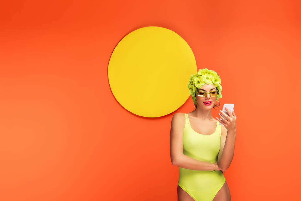 Mujer en sombrero con flores decorativas mirando el teléfono inteligente con círculo amarillo detrás sobre fondo naranja
 - Foto, Imagen