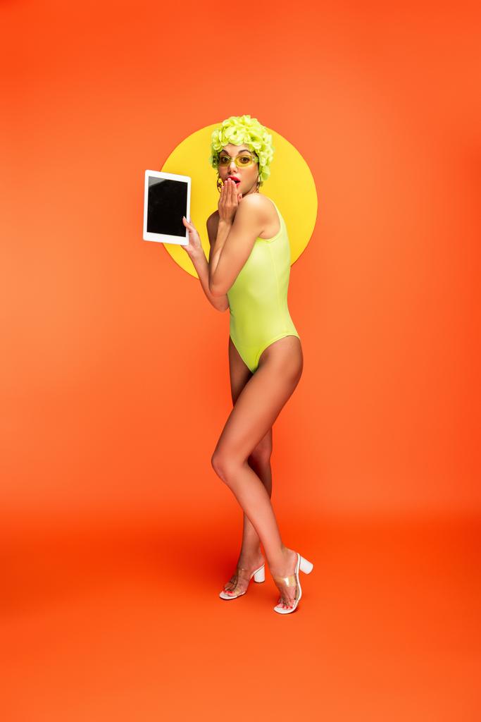 Schockierte Frau blickt in Kamera und zeigt digitales Tablet mit gelbem Kreis auf orange - Foto, Bild