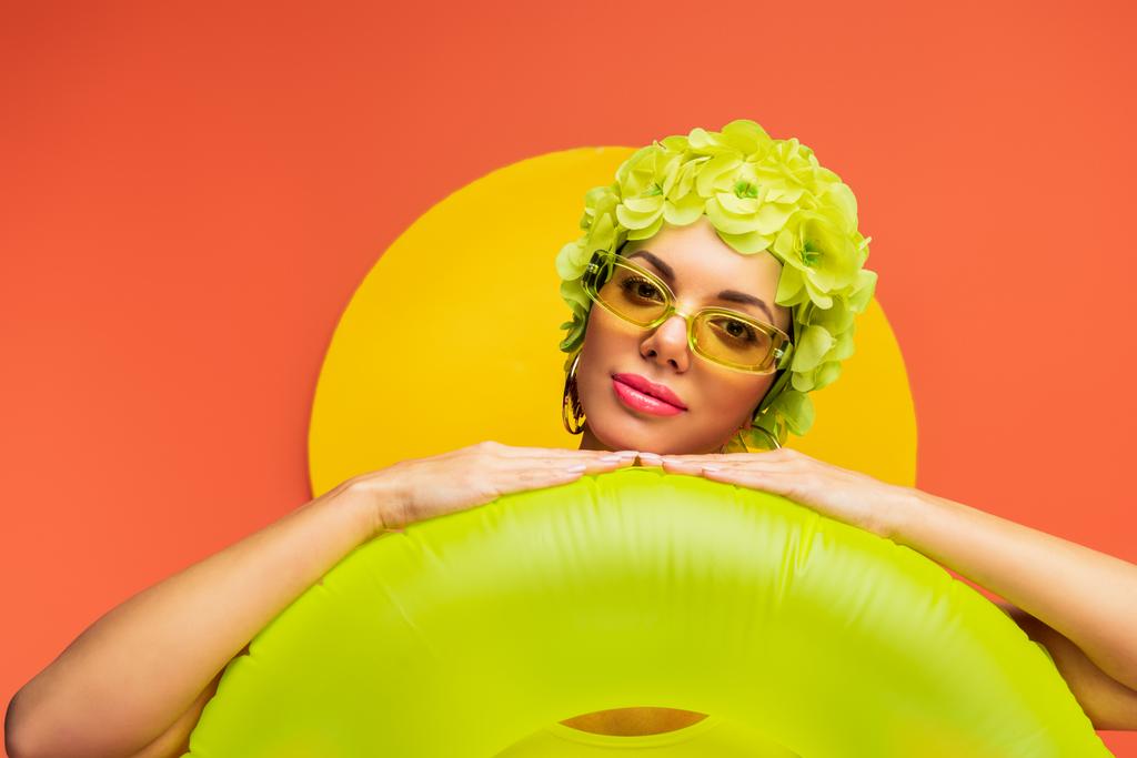 Portrait of girl in hat with decorative flowers and sunglasses putting hands on swim ring on yellow and orange  - Photo, Image