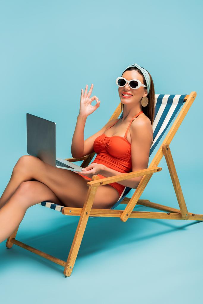 Freelancer met gekruiste benen en laptop glimlachend en met OK teken op ligstoel op blauw - Foto, afbeelding