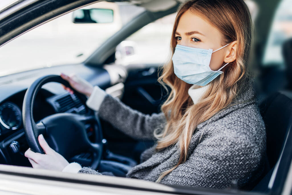 Красивая молодая девушка в маске сидит в машине, защитная маска от коронавируса, водитель на городской улице во время вспышки коронавируса, ковид-19 - Фото, изображение