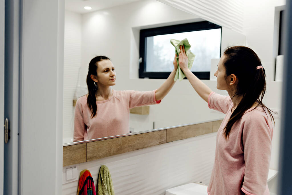 jong huisvrouw wast een spiegel met wasmiddel, gelukkig vrouw schoonmaken spiegel met doek thuis, mooi vrouw reinigt spiegel in de badkamer thuis, schoonmaken van de badkamer, spiegel schoonmaken - Foto, afbeelding
