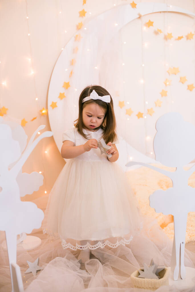 Cute little girl w białej sukience księżniczki z łukiem na włosach bawi się drewnianymi balerinami wśród złotych gwiazd na tle dużego białego drewnianego księżyca i girlandy. Wystrój dziecięcy. Urlop dla dzieci - Zdjęcie, obraz