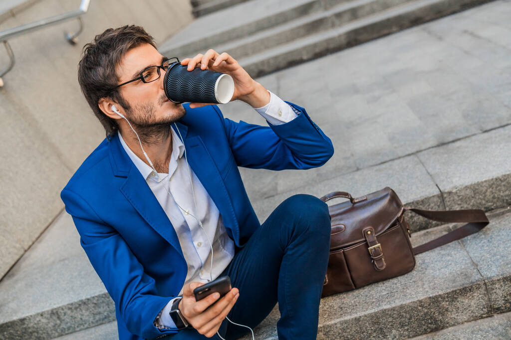 Nuori liikemies kuuntelee musiikkia älypuhelimesta juodessaan kahvia ulkona kaupungissa
 - Valokuva, kuva