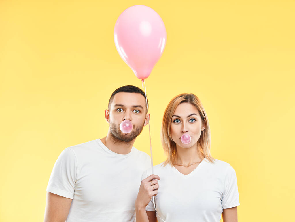 Νεαρό ζευγάρι φυσάει φούσκα με τσίχλα και κρατώντας ροζ μπαλόνι αέρα σε κίτρινο φόντο - Φωτογραφία, εικόνα