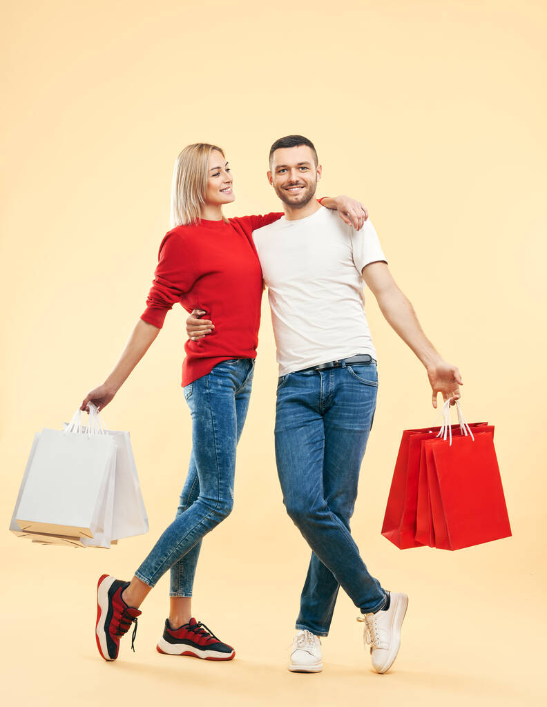 Plně dlouhý portrét mladého šťastného páru s nákupními taškami v rukou pózujících na béžovém pozadí. prodej, nákupní koncept - Fotografie, Obrázek