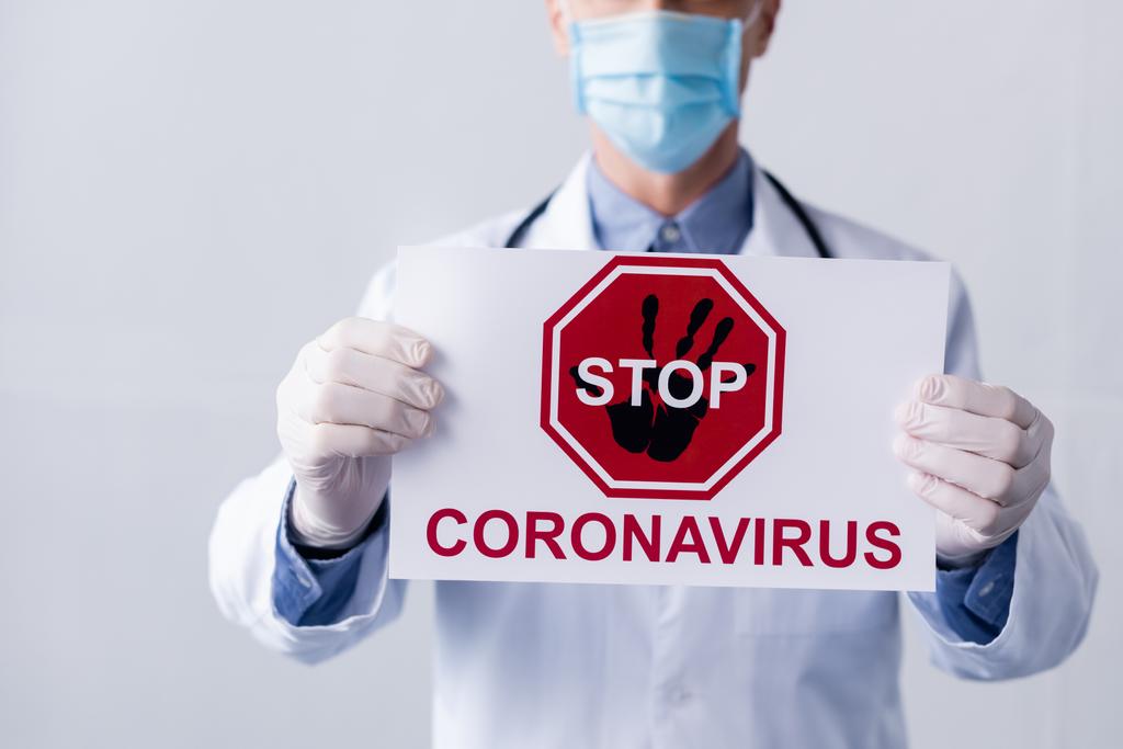 oříznutý pohled na zralého lékaře v bílém plášti a lékařské masce s nápisem stop koronaviru na šedé - Fotografie, Obrázek
