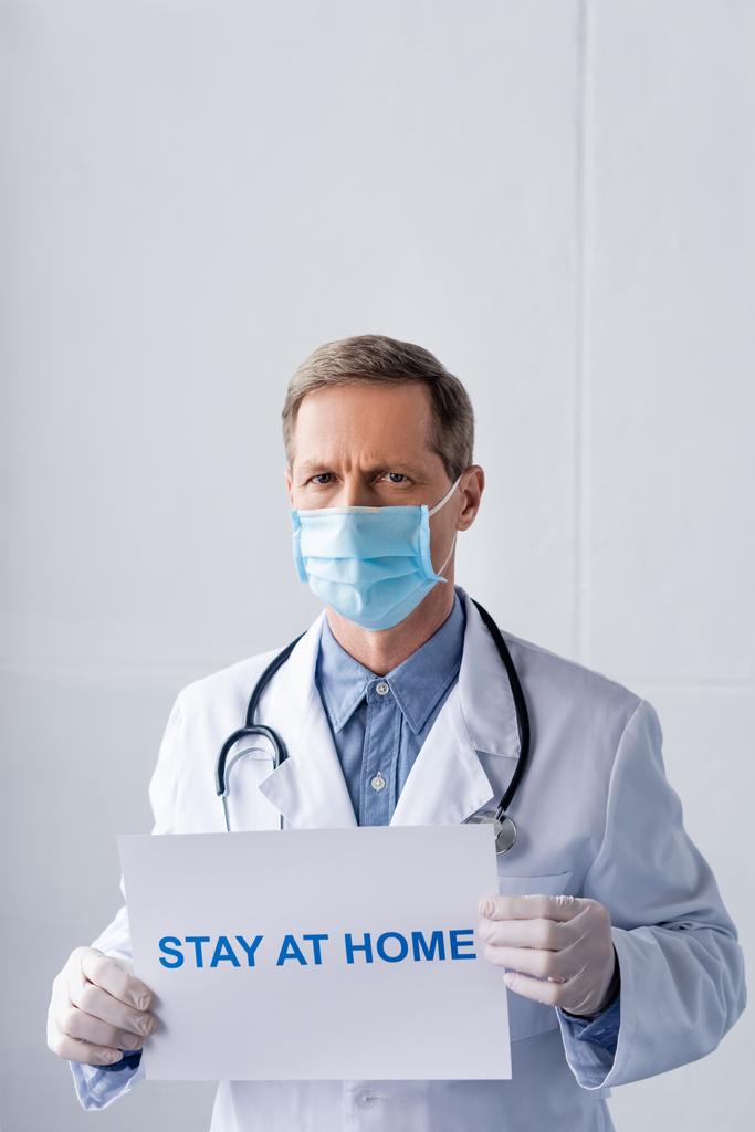 Arzt mittleren Alters in medizinischer Maske mit Plakat mit dem Schriftzug "zu Hause bleiben" auf grau - Foto, Bild