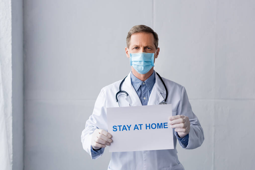 Dojrzały lekarz w masce medycznej i rękawiczkach lateksowych z tabliczką z napisem na szarym - Zdjęcie, obraz