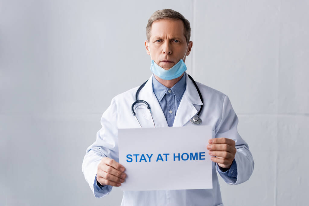Reifer Arzt in medizinischer Maske mit Plakat mit Bleibe-zu-Hause-Schriftzug auf grau - Foto, Bild