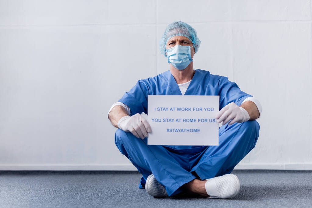 mature doctor in medical mask and cap holding placard with i stay at work for you, you stay at home for us lettering while sitting on white  - Photo, Image
