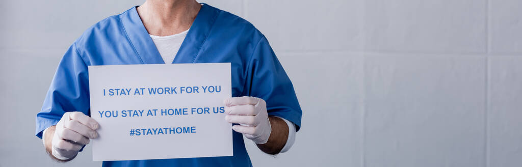 Panoramaaufnahme eines reifen Arztes mit Plakat "Ich bleib für dich bei der Arbeit, du bleibst für uns zu Hause" Schriftzug auf grau - Foto, Bild