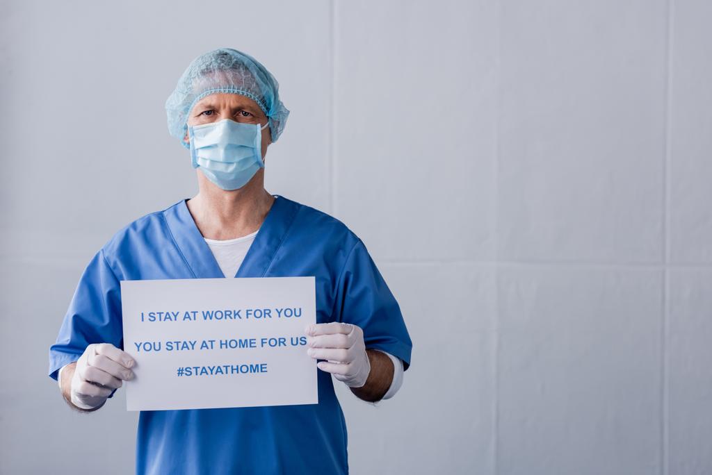 mature doctor in medical mask and cap holding board with i stay at work for you, you stay at home for us lettering on grey - Photo, Image