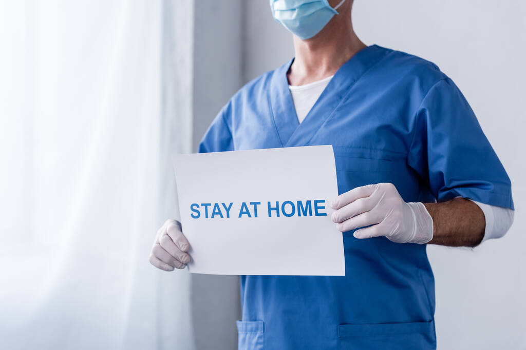 vista recortada del médico de mediana edad en la máscara médica con pancarta con letras de estancia en casa
  - Foto, imagen