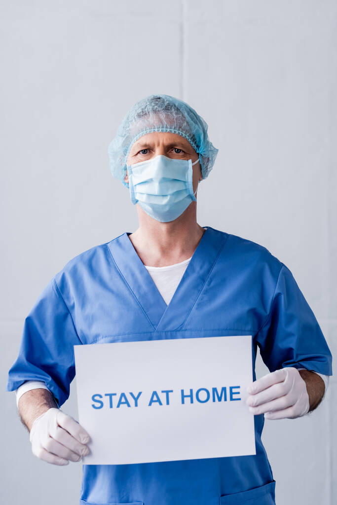 docteur mature en masque médical et casquette tenant la plaque avec le séjour à la maison lettrage sur gris
 - Photo, image