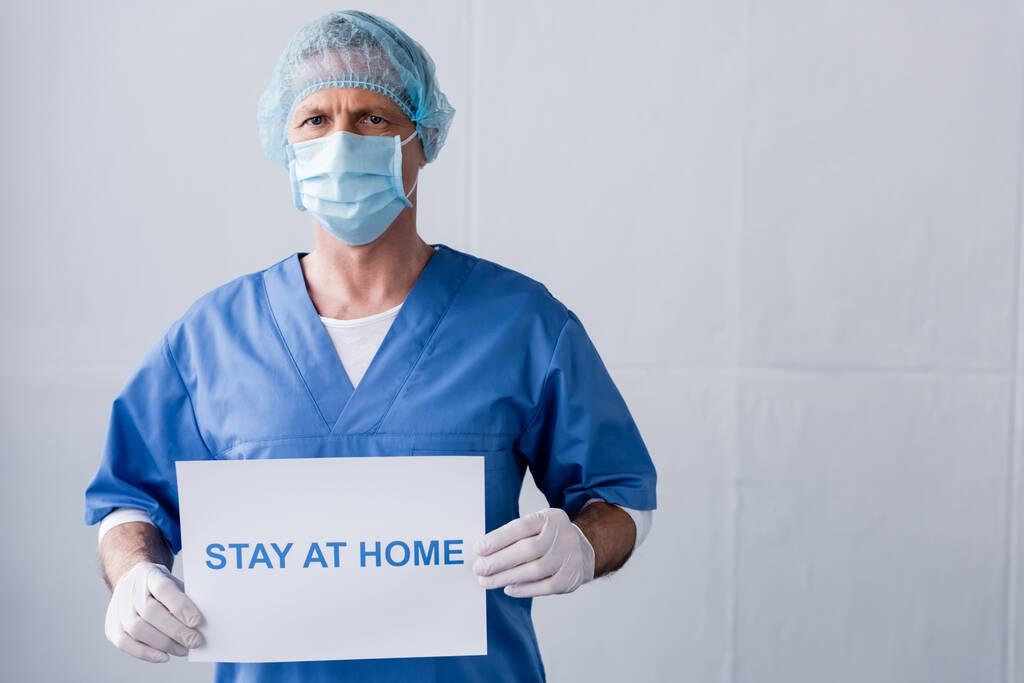 médecin mature en masque médical, bonnet et gants en latex tenant une pancarte avec inscription de séjour à la maison sur gris
 - Photo, image