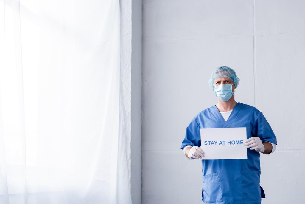 médico maduro em máscara médica, tampa e luvas de látex segurando cartaz com estadia em casa lettering
  - Foto, Imagem