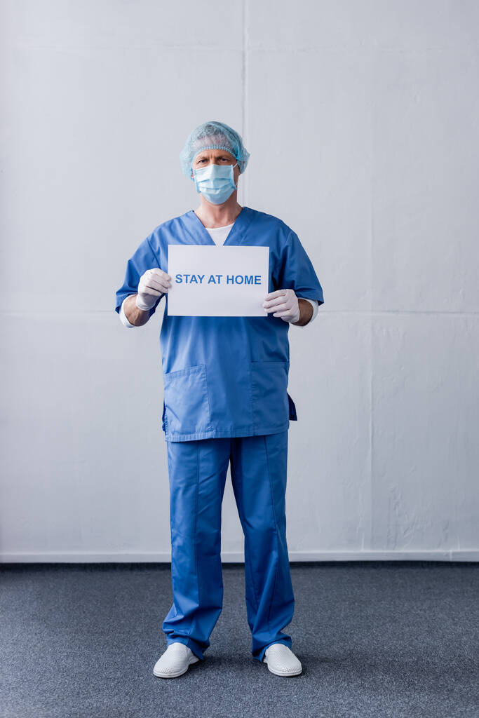 volwassen arts in medisch masker, pet en latex handschoenen met plakkaat met verblijf thuis belettering terwijl staan op grijs - Foto, afbeelding