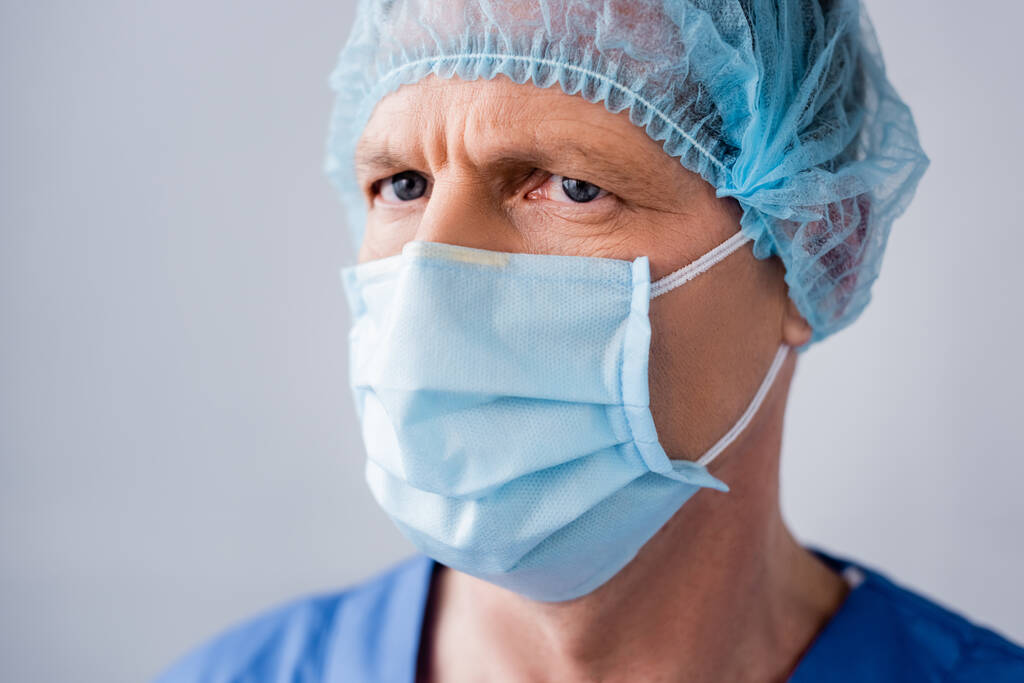 Dojrzały lekarz w niebieskiej masce medycznej i czapce izolowane na szarości  - Zdjęcie, obraz