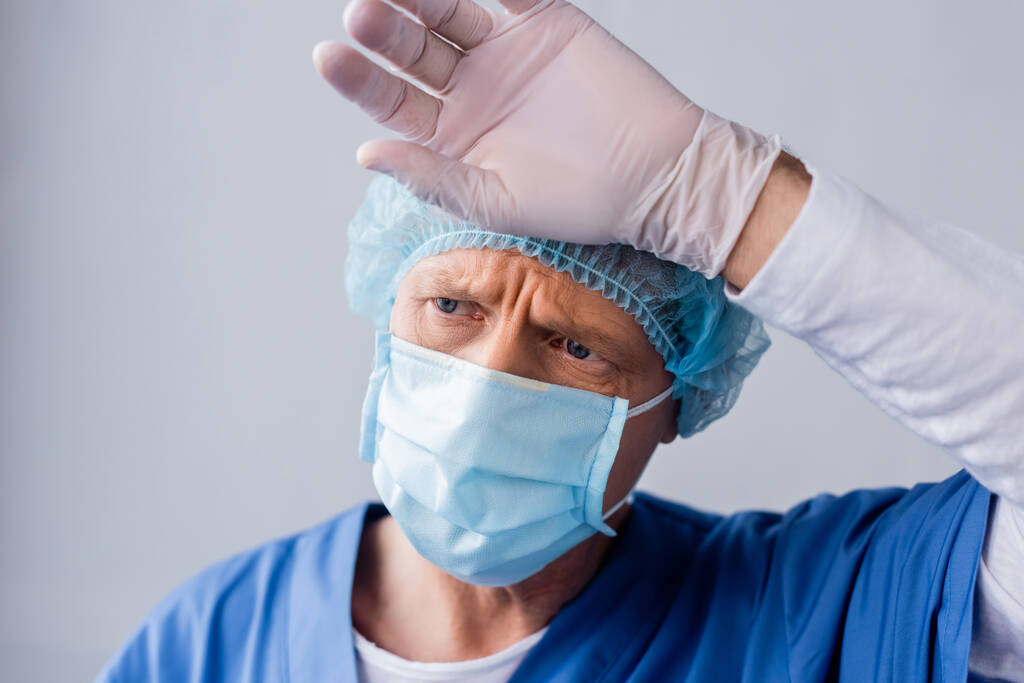 κουρασμένος και ώριμος γιατρός σε μπλε ιατρική μάσκα και καπάκι αγγίζοντας το μέτωπο σε γκρι  - Φωτογραφία, εικόνα