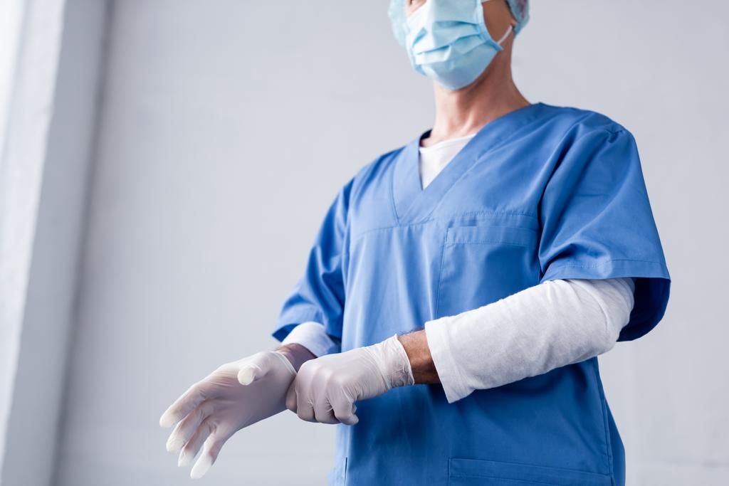 καλλιεργημένη άποψη του ώριμου γιατρού σε μπλε ιατρική μάσκα φορώντας γάντι λατέξ σε γκρι - Φωτογραφία, εικόνα