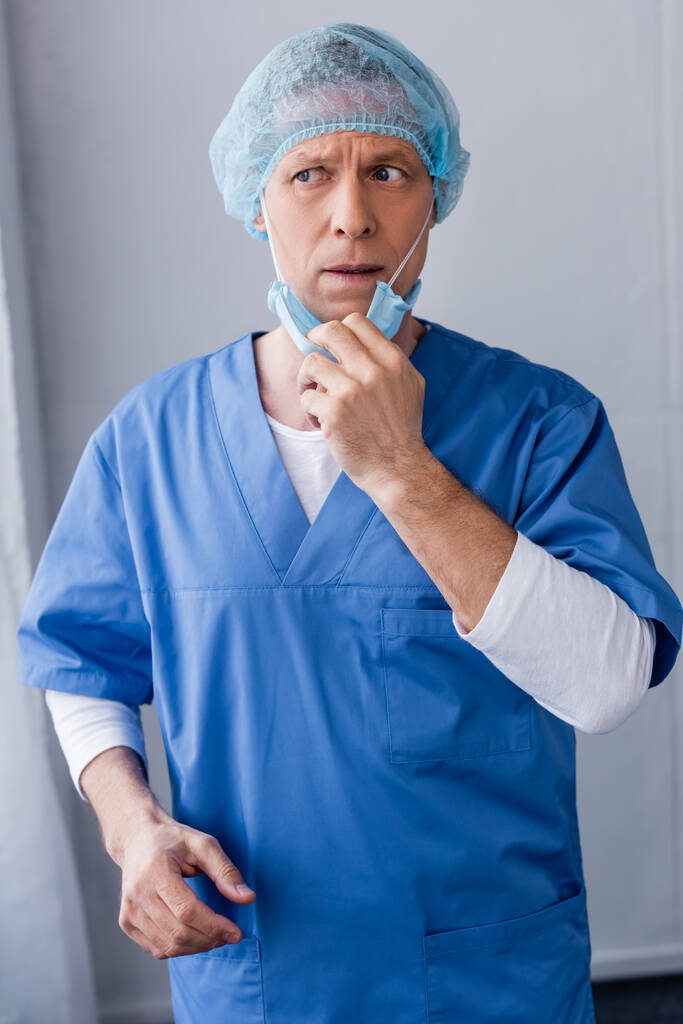 mature doctor in blue medical cap touching medical mask and looking away  - Photo, Image