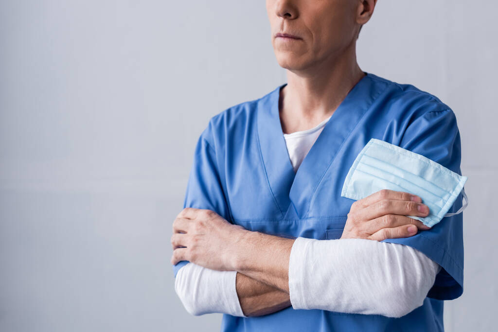 oříznutý pohled na lékaře středního věku stojícího se zkříženýma rukama a držícího lékařskou masku na šedé - Fotografie, Obrázek