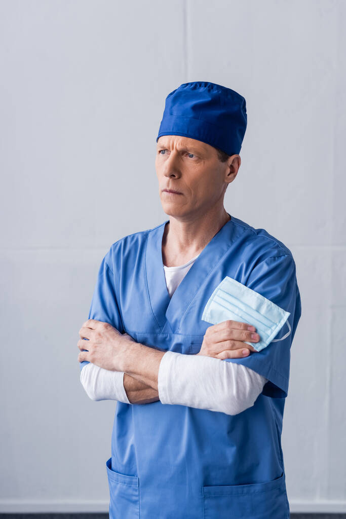 arts van middelbare leeftijd in blauwe scrub hoed staan met gekruiste armen en het houden van medische masker op grijs - Foto, afbeelding
