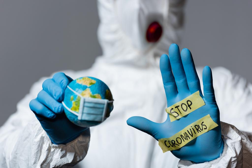 selective focus of man in hazmat suit holding globe and showing hand with stop coronavirus lettering isolated on grey  - Photo, Image