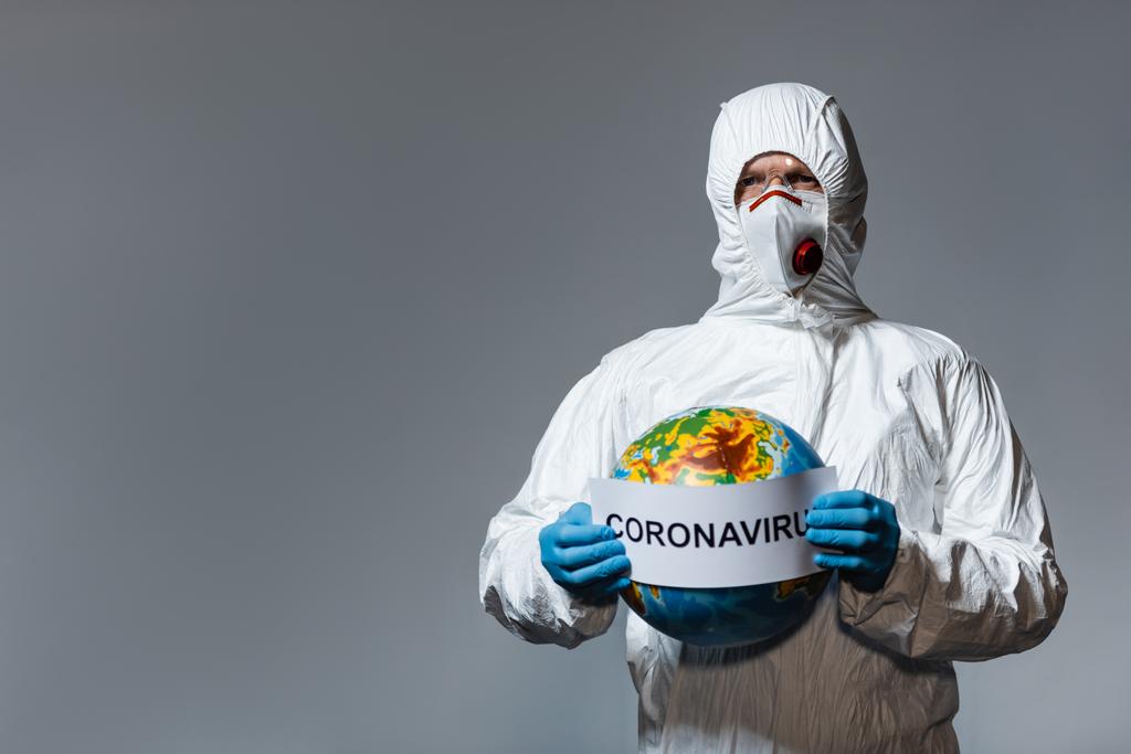 uomo in tuta hazmat e maschera medica che tiene globo e carta con scritte coronavirus isolate su grigio
  - Foto, immagini
