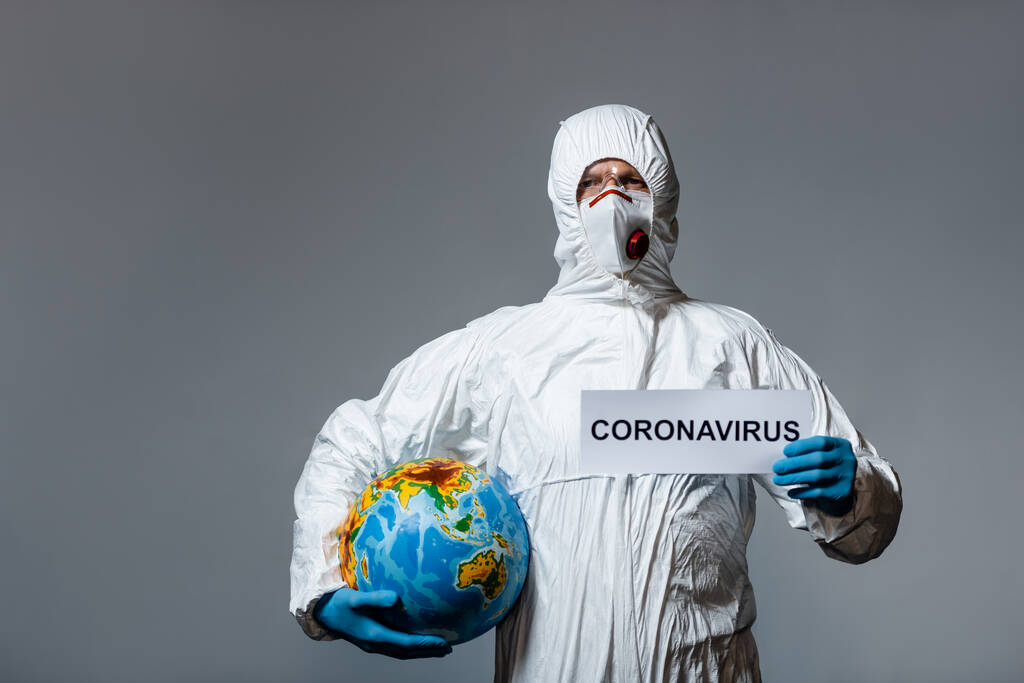 mens in persoonlijke beschermingsmiddelen met wereldbol en papier met coronavirusbelettering geïsoleerd op grijs  - Foto, afbeelding