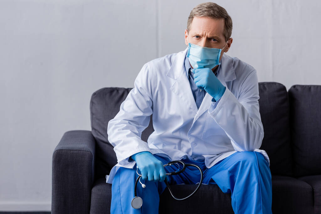 médico maduro em máscara médica segurando estetoscópio enquanto sentado no sofá
  - Foto, Imagem