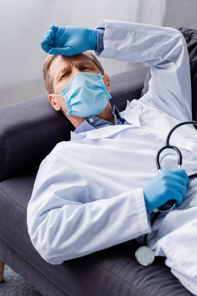 zmęczony i dojrzały lekarz w masce medycznej trzymając stetoskop leżąc na sofie  - Zdjęcie, obraz