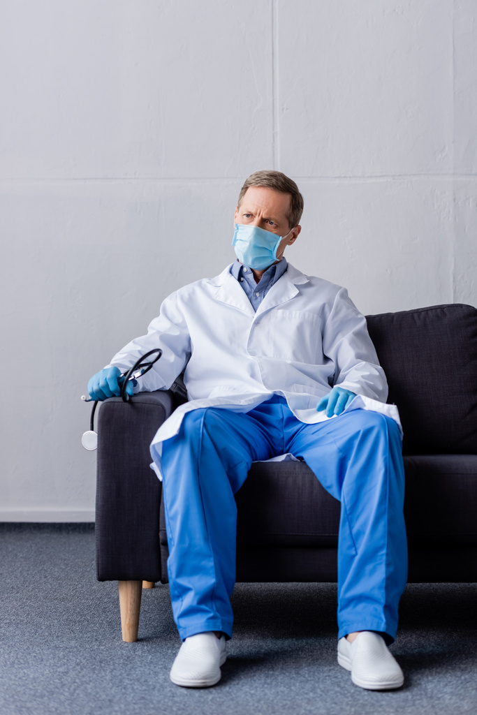 volwassen arts in medisch masker houden stethoscoop terwijl zitten op de bank  - Foto, afbeelding