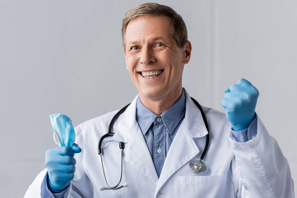 счастливый и зрелый доктор в латексных перчатках держит медицинскую маску на сером
 - Фото, изображение