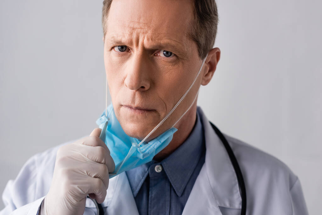 zralé lékař v latexové rukavici dotýkající se modré lékařské masky na šedé  - Fotografie, Obrázek