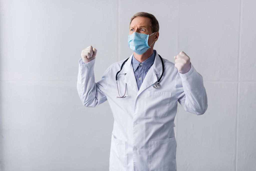 Stresli orta yaşlı doktor mavi tıbbi maskeli Gri yumruklarıyla ayakta duruyor  - Fotoğraf, Görsel