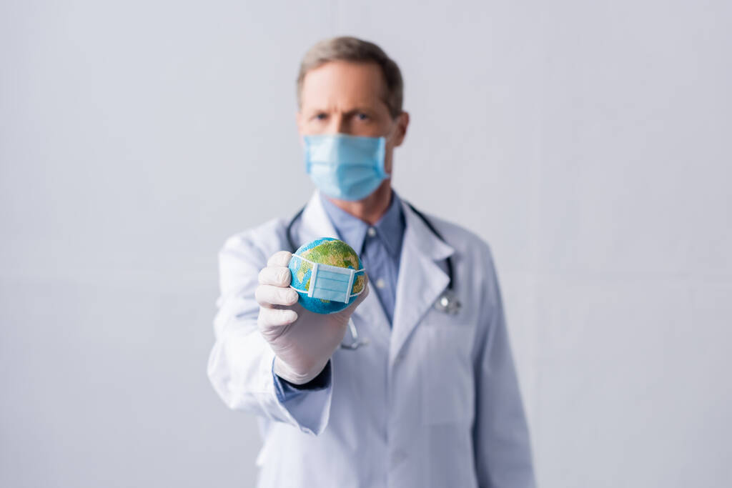 selektivní zaměření lékaře středního věku v latexové rukavici a lékařské masce držící malý glóbus na šedé - Fotografie, Obrázek