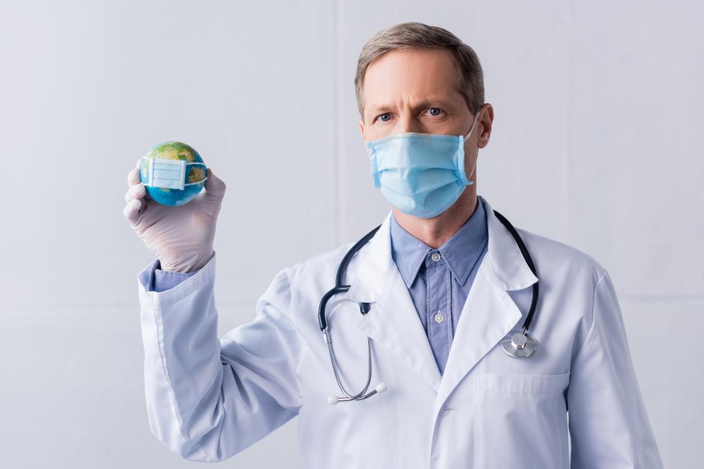 Dojrzały lekarz w lateksowej rękawiczce i masce medycznej trzymający mały glob na szarości - Zdjęcie, obraz