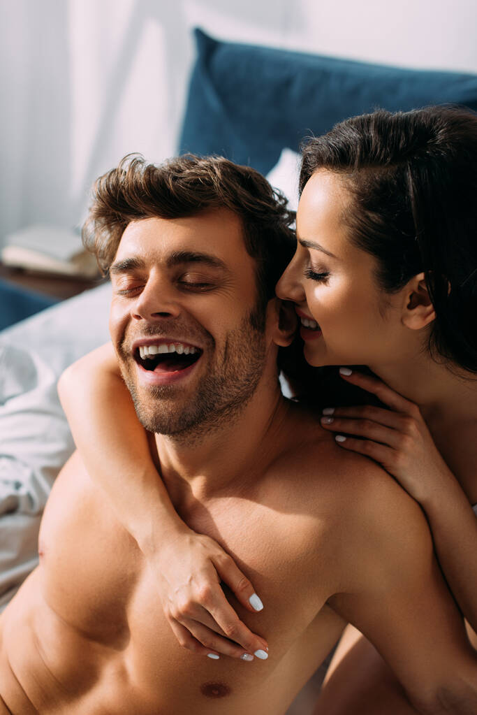 Γυναίκα με κλειστά μάτια χαμογελώντας και αγκαλιάζοντας ενθουσιασμένο φίλο από πίσω στο κρεβάτι στην κρεβατοκάμαρα - Φωτογραφία, εικόνα