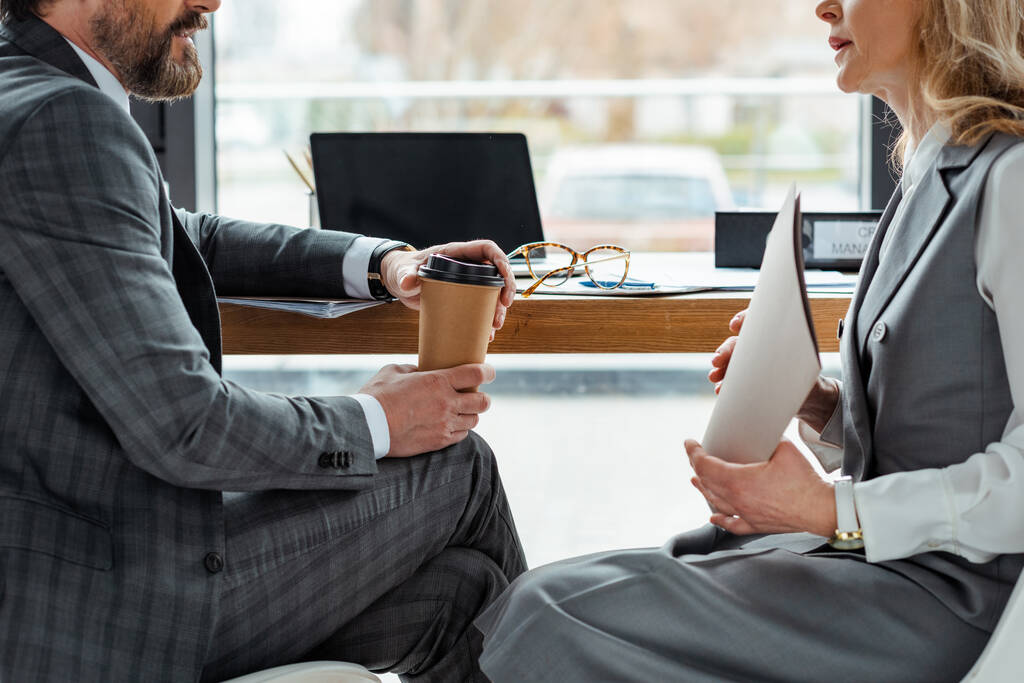 Przycięty widok biznesmenów z kawą na wynos i teczką papierową siedzącą przy stole w biurze  - Zdjęcie, obraz