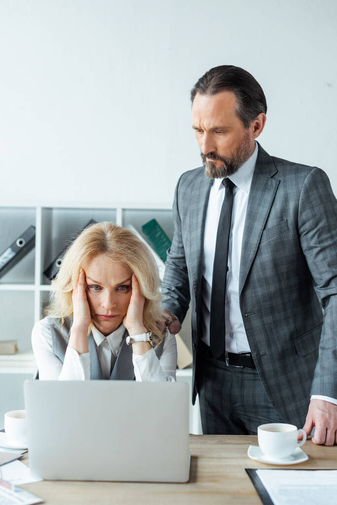Красивий бізнесмен стоїть біля концентрованої бізнес-леді, дивлячись на ноутбук в офісі
  - Фото, зображення