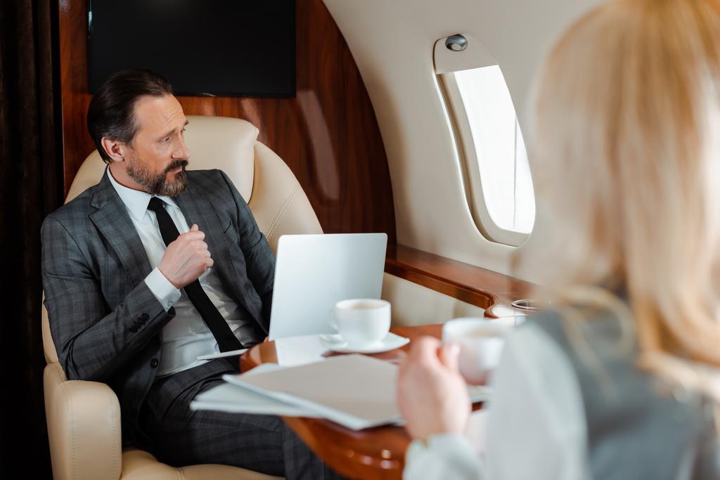 ビジネスの近くに座っているノートパソコンを持つビジネスマンの選択的な焦点飛行機の中でコーヒーを飲みながら  - 写真・画像