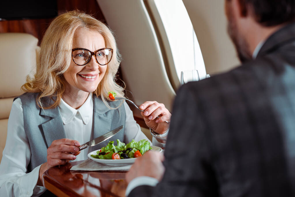 Selektywne skupienie uśmiechniętej bizneswoman jedzącej sałatkę w pobliżu biznesmena w samolocie  - Zdjęcie, obraz