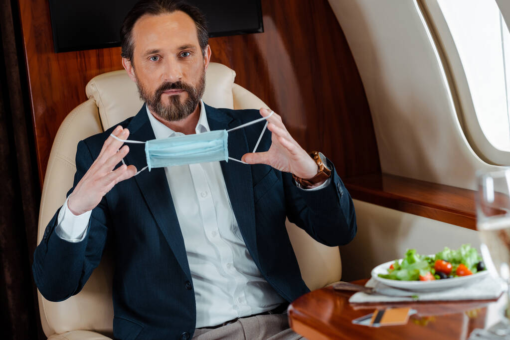Selective focus of businessman holding medical mask near salad, glass of champagne and credit cards on table in plane  - Photo, Image