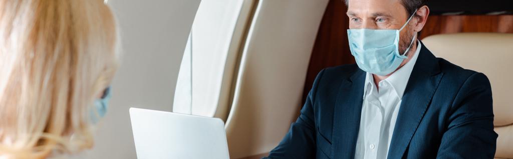 Foco seletivo de homem de negócios em máscara médica usando computador portátil perto de empresária em avião, tiro panorâmico
  - Foto, Imagem