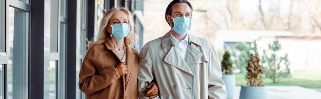 Panoramatický záběr obchodní pár v lékařských maskách při pohledu pryč v blízkosti budovy na městské ulici  - Fotografie, Obrázek