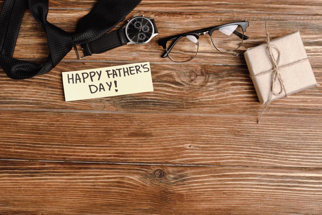 Bovenaanzicht van geschenkdoos, wenskaart met belettering gelukkige vaderdag en heren zwarte das, polshorloge en bril op houten achtergrond - Foto, afbeelding