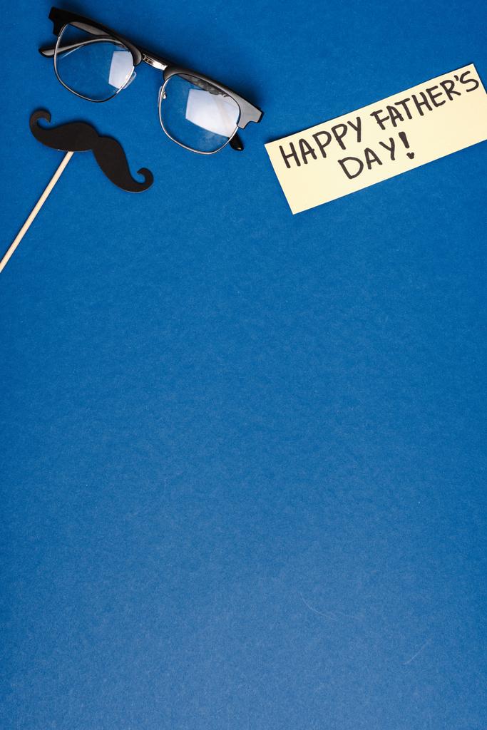 Draufsicht auf Herrenbrille, Schmuckkarton falscher Schnurrbart und Grußkarte mit Schriftzug Happy Vatertag auf blauem Hintergrund - Foto, Bild