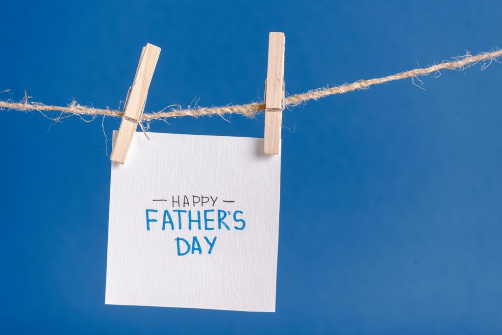 Papel branco cartão de saudação com letras escritas à mão feliz dia dos pais pendurado na corda com varas isoladas em azul
 - Foto, Imagem